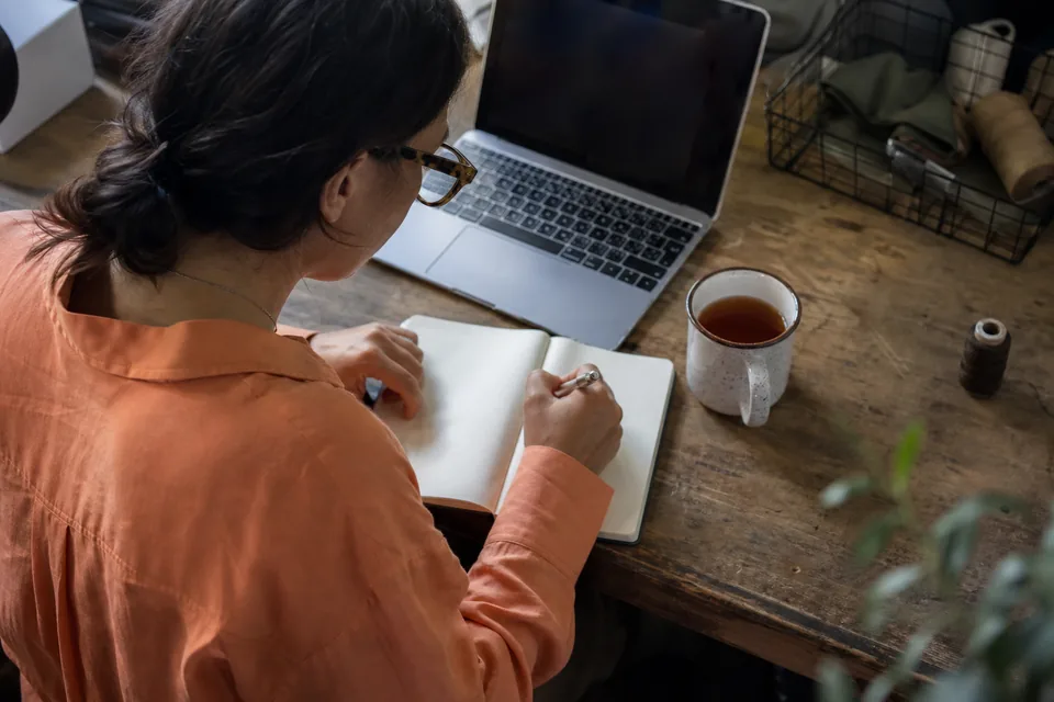 A creator writing price ideas in a notebook next to a laptop and a cup of tea
